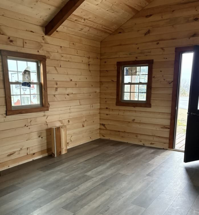Small cabin interior with light pine wood paneling, gray laminate flooring, and natural light from windows